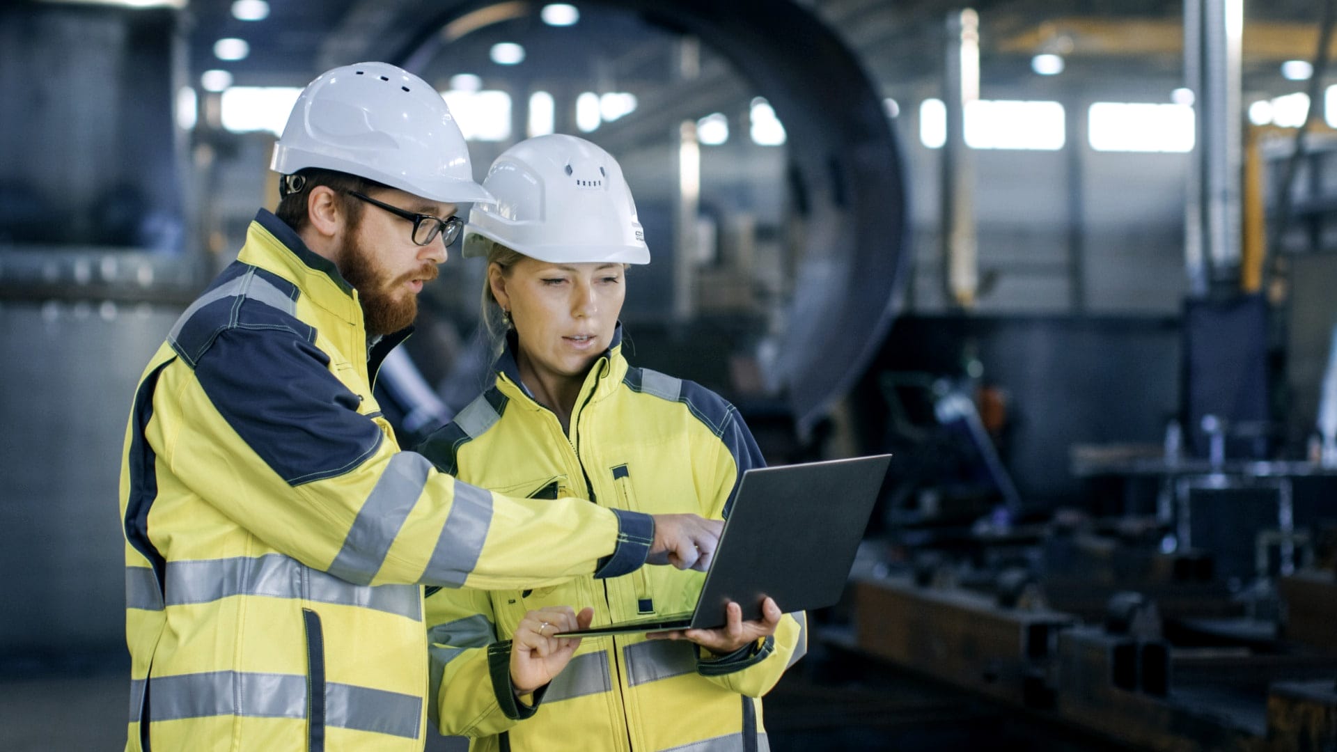 Two engineers in hard hats and high-visibility jackets review a tablet in an industrial plant.