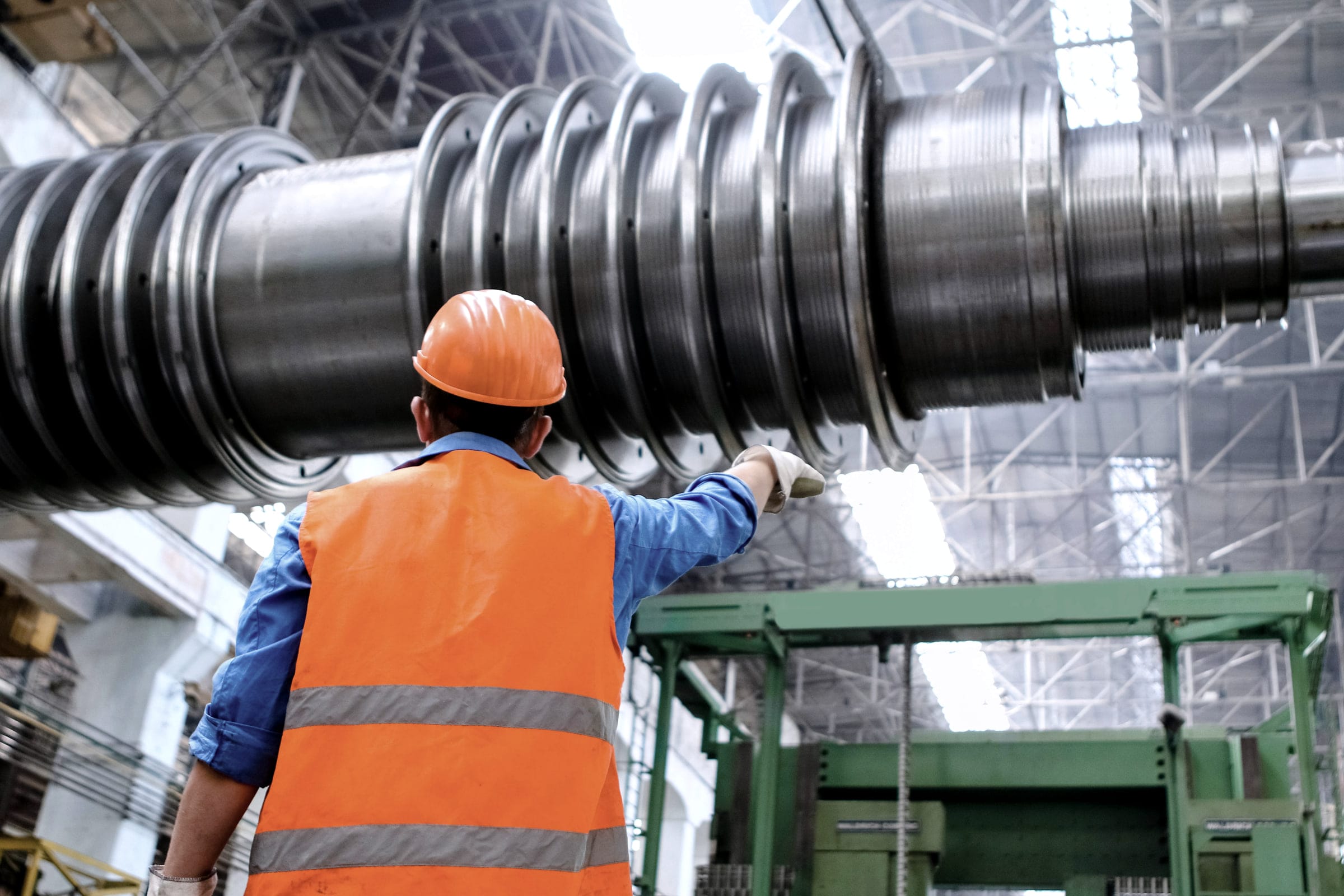 A worker in a hard hat and high-visibility vest inspects a large machined shaft.