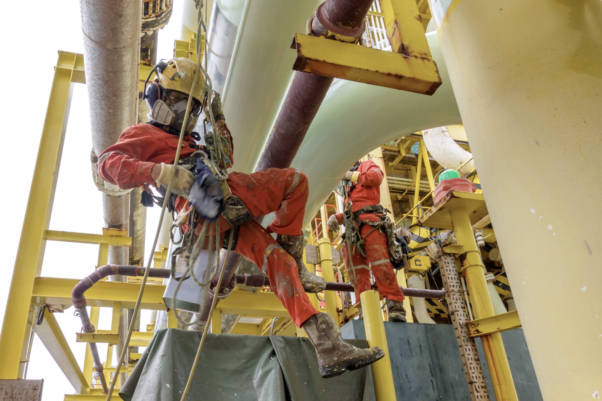Rope-access technicians in red coveralls working on a yellow offshore structure.